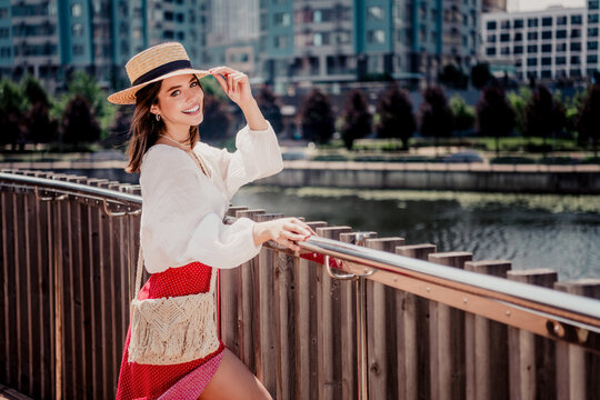 Smiling young woman in a straw hat and red dress leaning on a waterfront railing in an urban summer setting