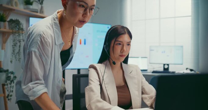Young Asian businesswoman mentoring trainee in call center office. Corporate customer service training, employee support coach, team communication help desk, telemarketing agent work concept.