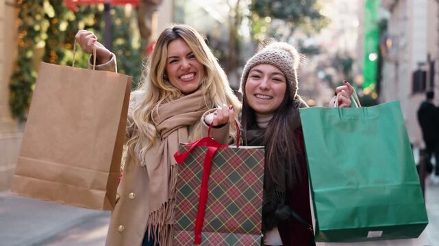 Video of two happy female friends showing the shopping bags while looking at the camera on the city streets in winter.