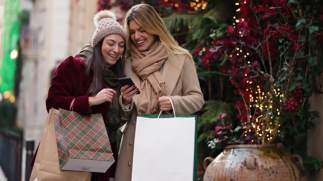 Video of two beautiful friends using smartphone while walking on the city street with shopping bags.