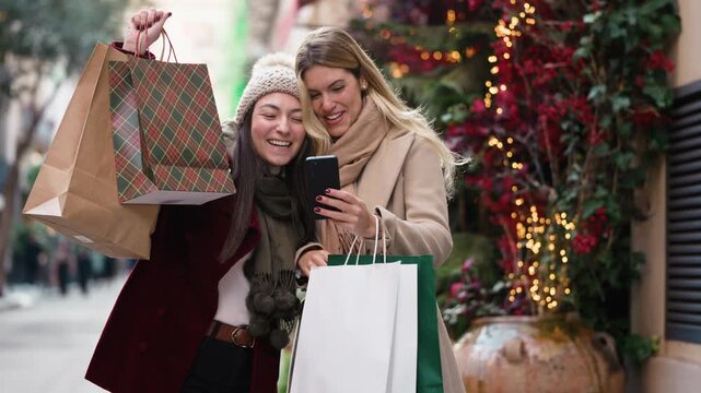 Video of two beautiful friends doing videocall with smartphone while walking on the city street with shopping bags.