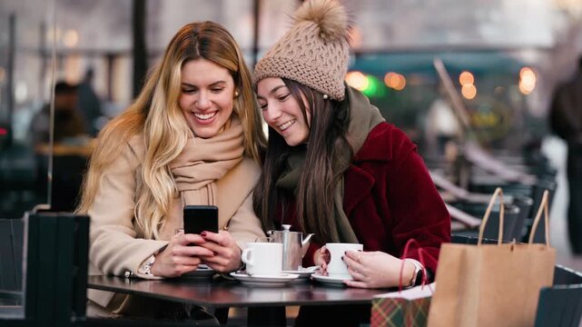Video of two beautiful women using smartphone while drinking coffee sitting in the bar terrace.