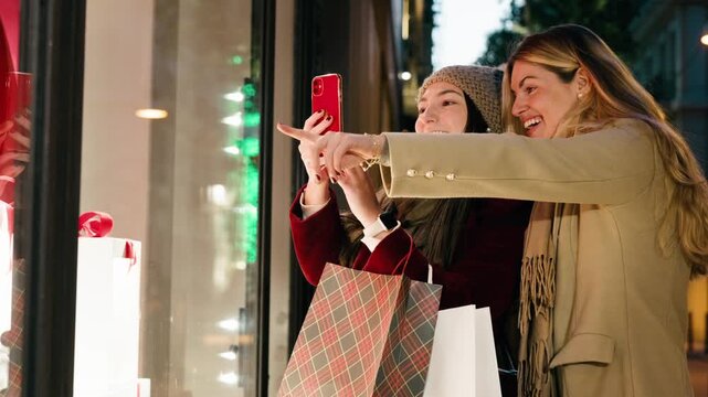 Video of two beautiful women taking a photo with smartphone of a shop window at night in winter.