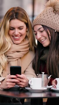 Video of two beautiful women using smartphone while drinking coffee sitting in the bar terrace.