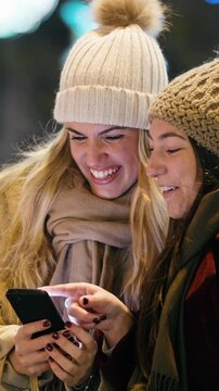 Video of two beautiful friends using smartphone while walking on the city street with shopping bag.