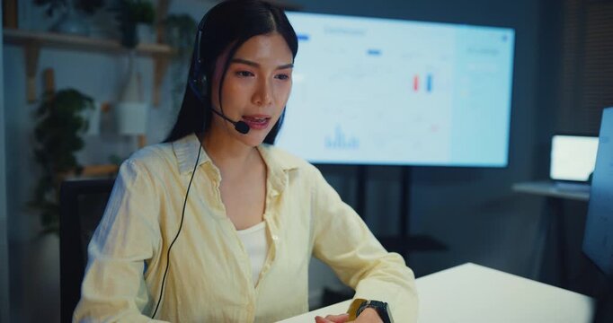 Young Asian businesswoman working late talk on telephone headset in call center office at night. Corporate customer service support, telemarketing agent, finance insurance investment advisor concept.