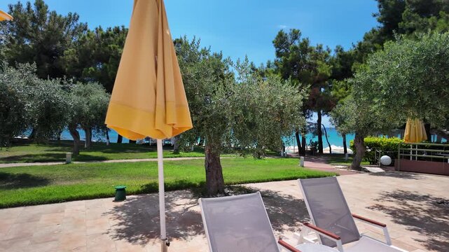 The terrace of a luxury island hotel overlooks the turquoise Aegean Sea, a green garden with olive trees, and a private beach in sunny Greece