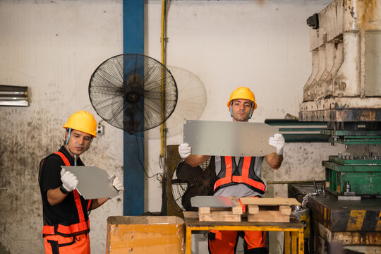 Two machine operators each hold up a stamped sheet metal panel to inspect against the light beside a power press machine