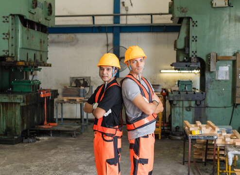 Two male workers in orange vests and hard hats stand back to back, arms crossed, between power presses on factory floor