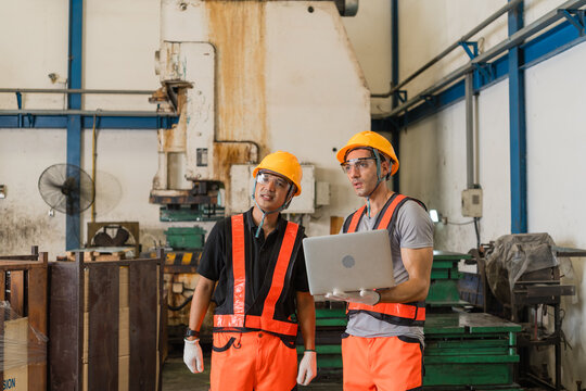 Close portrait of factory engineers reviewing laptop records near aging power press machine supporting efficient production and workplace safety inside a busy metalworking factory setting