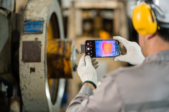 Engineer in yellow ear protection using thermal imaging to photograph a power press flywheel with a smartphone for predictive maintenance