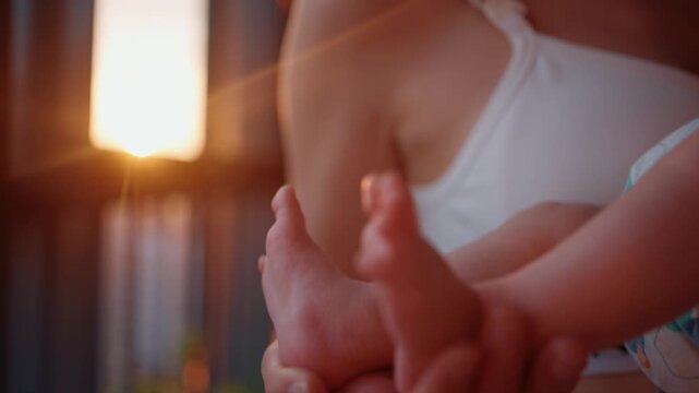 Close up view of a mother's hands tenderly holding and caressing the small, delicate feet of her newborn child, with a beautiful golden sunset flare in the background