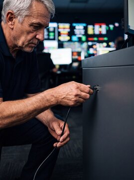 Nahaufnahme der H&auml;nde eines &auml;lteren Technikers der ein Datenkabel an eine Konsole in einer Einsatzzentrale bei Nacht anschlie&szlig;t