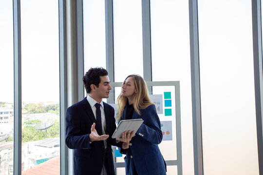 Two business professionals discussing marketing strategy and business analytics in front of a glass board with charts and sticky notes.Corporate colleagues collaborating on project planning,