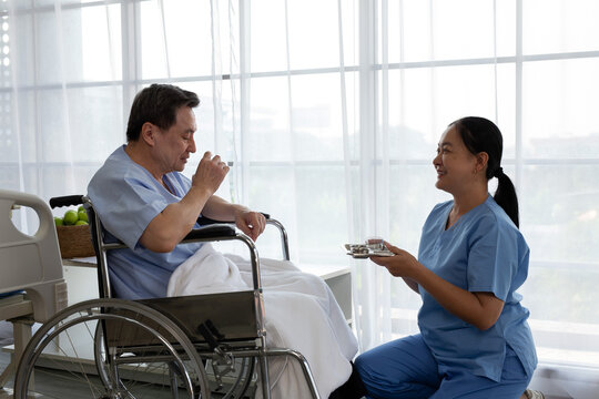 Smiling nurse giving medication and a glass of water to an elderly patient sitting in a wheelchair in a hospital room. Senior healthcare, nursing care, rehabilitation, compassion