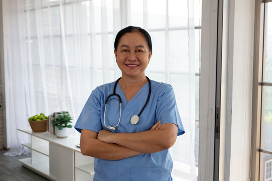 Portrait of smiling mature Asian woman doctor in blue scrubs. Confident middle-aged  medical professional in blue uniform with stethoscope, crossed arms.