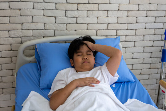 Sick young boy lying in a hospital bed with his hand on his forehead, resting during medical treatment and recovery. Healthcare, pediatric care, illness, fever, patient care, and hospital concept.