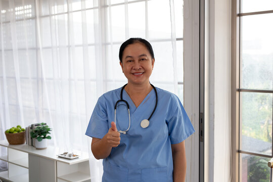 Portrait of smiling mature Asian woman doctor in blue scrubs. Confident middle-aged  medical professional in blue uniform with stethoscope, crossed arms.