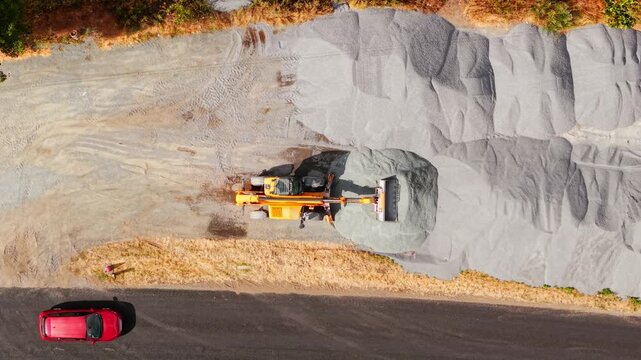 Construction excavator road repair. Parked telehandler beside crushed stone aggregate stockpile with aerial top view. Highway maintenance equipment and infrastructure materials along asphalt roadside