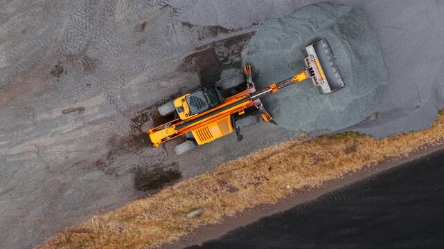 Construction tractor gravel stockpile. Parked telehandler beside crushed stone for road repair from aerial drone view. Highway infrastructure materials storage with heavy equipment and asphalt roadway
