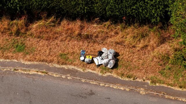 Illegal dumping roadside. Garbage bags discarded on grass verge beside asphalt road with aerial drone view. Fly tipping waste pollution near roadside vegetation and environmental contamination