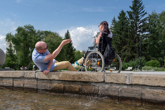 husband plays jokes with a paralyzed female disabled man in a wheelchair on a walk by the fountain splashing water