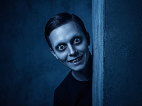 Creepy man with wide staring eyes and sinister grin peeking from behind a wall in dark blue lighting perfect for horror thriller and suspense projects