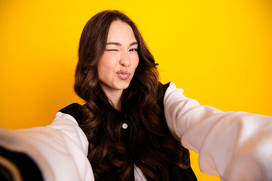 Young woman taking a playful selfie winking and puckering lips against a bright yellow background, casual portrait showing fun expression and confident modern style
