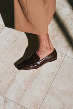 Close-up of a polished brown leather loafer worn with tailored beige trousers on sunlit stone tiles.