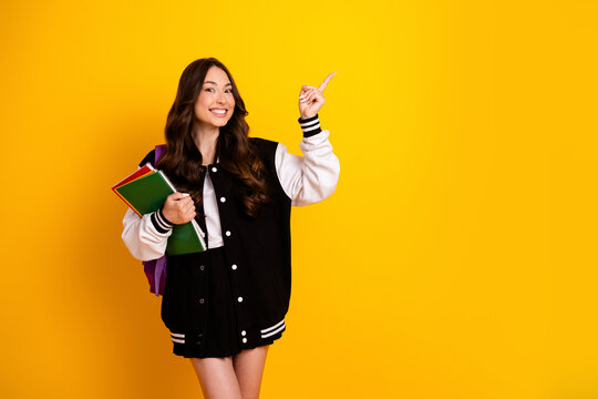 Smiling female student in a varsity jacket holding notebooks and backpack, pointing up with enthusiasm on a bright yellow background for school, college and education themes