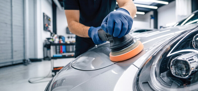 A person is working on a car in a garage. They hold a polishing machine and are moving it across the car's surface. Tools and equipment are arranged in the background
