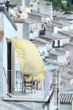 small balcony with yellow and white striped curtain billowing in the wind against the blurred backdrop of the houses of Leonforte, Sicily, Italy