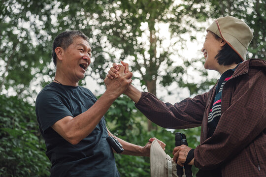 Elderly Asian couple laughing during joyful dap up hand pose with bright bokeh sunlight of forest trail, capturing romantic hiking and warmth nature connection activity for healthy ageing relationship