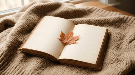 Fototapeta premium Open book with dry autumn leaf on beige knitted sweater. Cozy reading time at home. Still life featuring literature, seasonal foliage and textile texture for fall atmosphere concept.