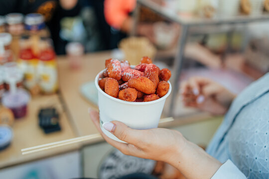Close-up of a hand holding a paper cup filled with golden mini fried doughnuts dusted with powdered sugar at a food stall