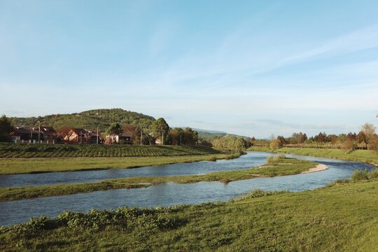A wide scenic shot of a winding river flowing through a lush green valley with rural houses nestled at the foot of a forested hill under a clear blue sky. Mukachevo city.