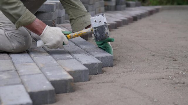 Construction worker positions gray paving block beside aligned row of pavers on outdoor walkway, gloved hand guiding placement near resting mallet, stacked blocks, sandy ground