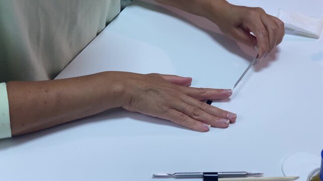 A calm woman gently files her fingernails with a slim metal file. She shapes her nails at a clean white table. This peaceful self care routine creates a relaxed mood.