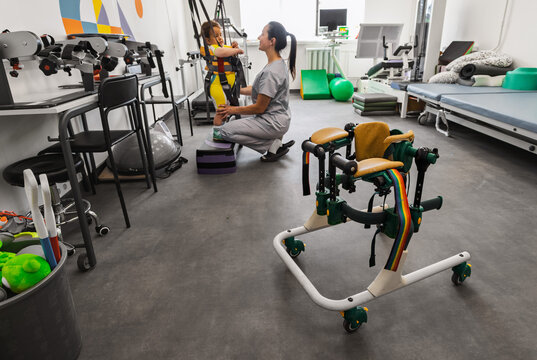 Children's walkers stand in rehabilitation room, in background a physiotherapist is doing rehabilitation exercises with little boy