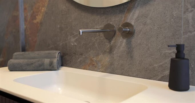 View of luxury modern bathroom with built in wall faucet and stone basin, slow camera panorama reveals dark natural stone cladding wooden vanity rounded mirror and dark accessories shaping interior