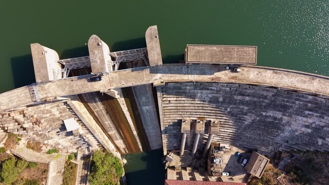 Top-down perspective of a concrete dam showing three sluice gates, stepped spillway face, turbine housing with machinery, and green reservoir water alongside the structure