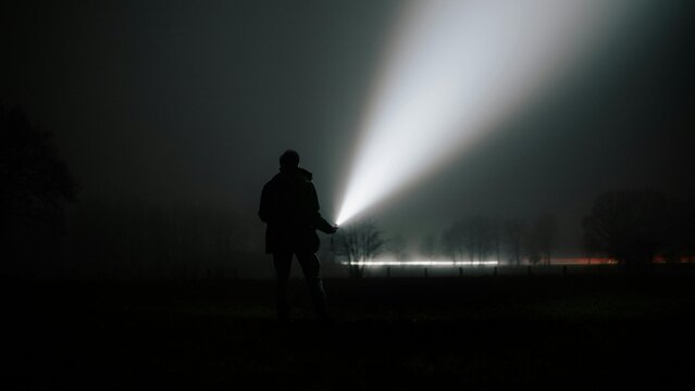 Silhouette of man holding flashlight emitting beam into dark foggy night landscape