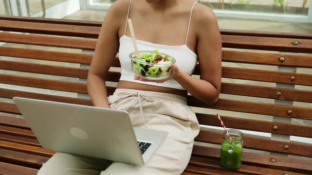 Young freelancer sitting on a bench, having lunch while working remotely on her computer