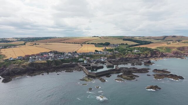 Aerial drone flight directly forward over the historic fishing village and harbour of St Abbs, revealing the scenic rugged Berwickshire coast and rural rolling fields under a cloudy sky in Scotland.