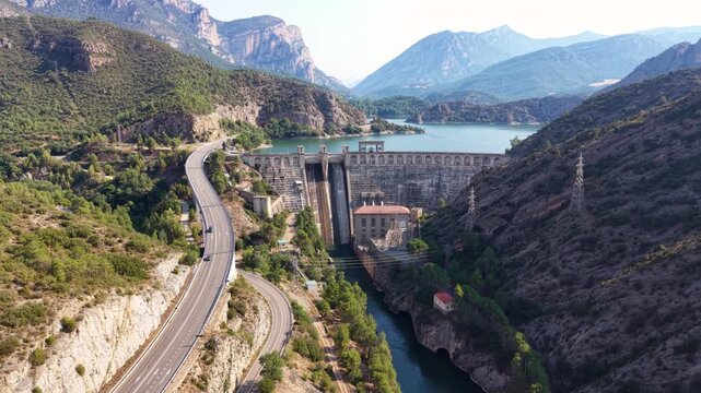 Curving highway winds along a rocky canyon toward a concrete gravity dam where a turquoise reservoir meets the structure amid layered mountain peaks in Lleida, Spain