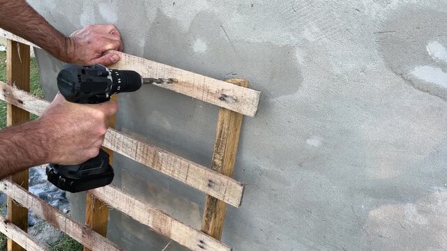Construction worker's hands drilling into a wooden pallet attached to a concrete wall using a modern cordless power tool for a diy home improvement project