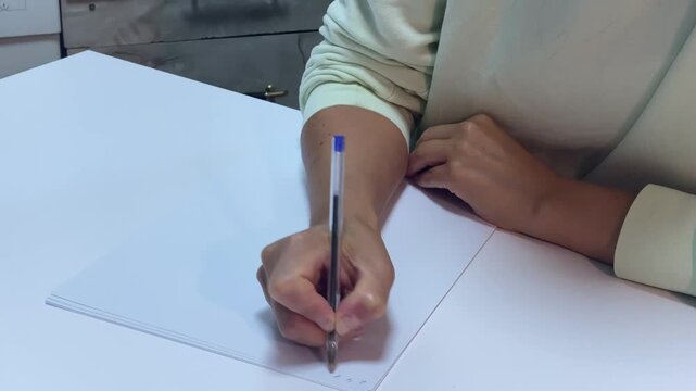A student holds a pen over blank paper to write. She rests quietly at a smooth white desk. Her calm presence brings a peaceful feeling.