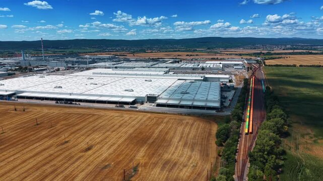 Factory and freight railway across rural landscape. White industrial halls border golden stubble and green crops beneath a vivid blue sky with cumulus clouds.