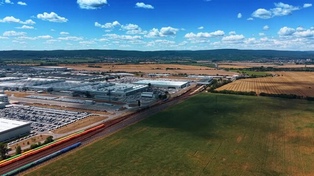 Factory vehicle yards beside freight railway. Rows of parked cars and white production halls border colorful railcars, fields, and wooded hills.