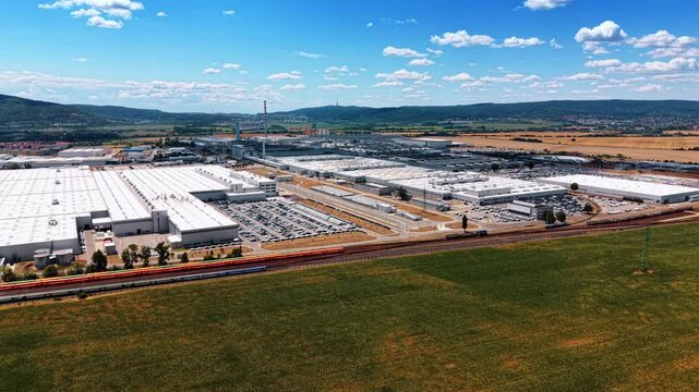 Manufacturing complex behind railway and green field. Multiple white halls and vehicle yards spread across a valley beneath scattered clouds and wooded ridges.